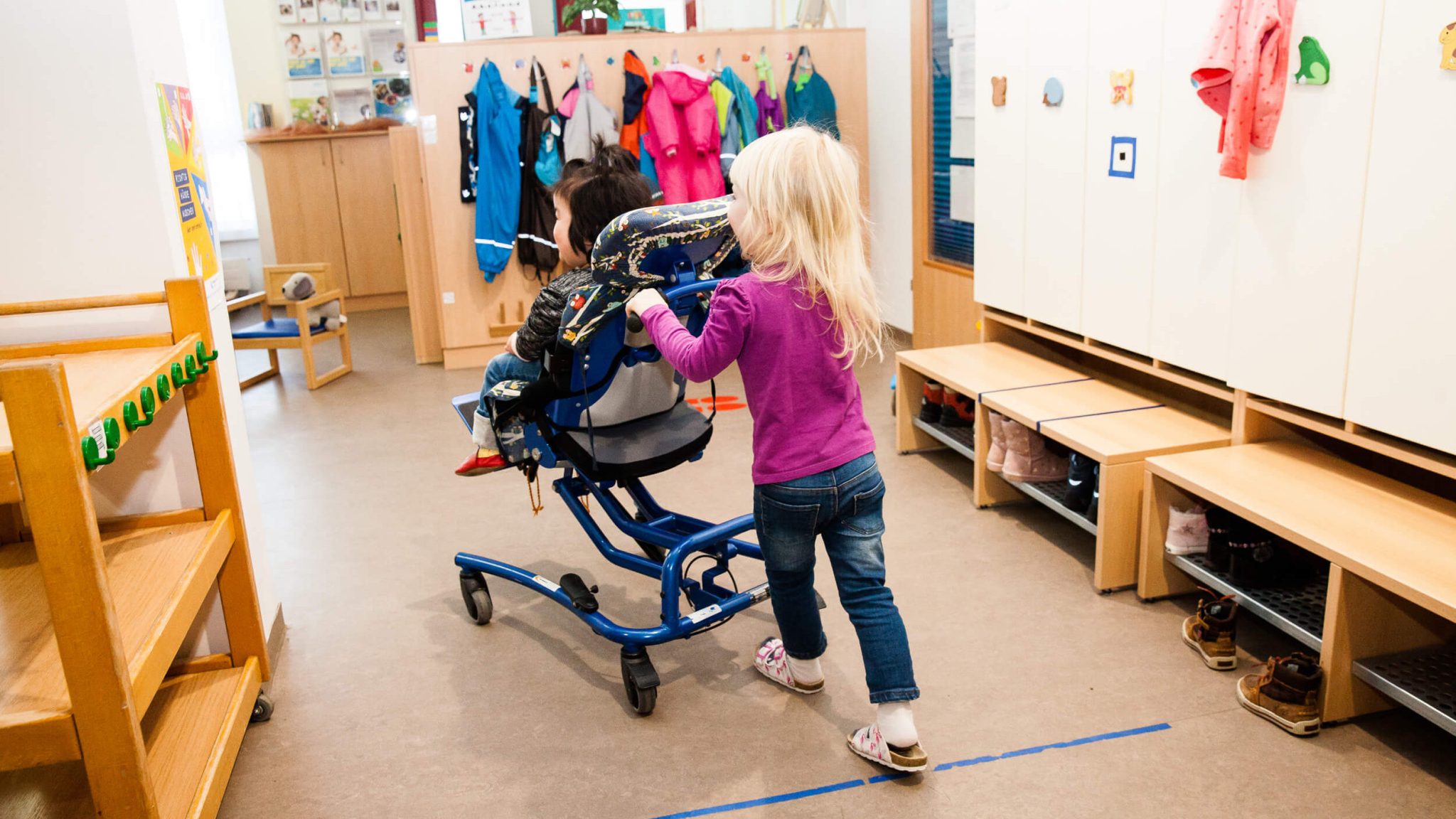 Ein Mädchen schiebt ein Mädchen im Rollstuhl durch einen Kindergarten.